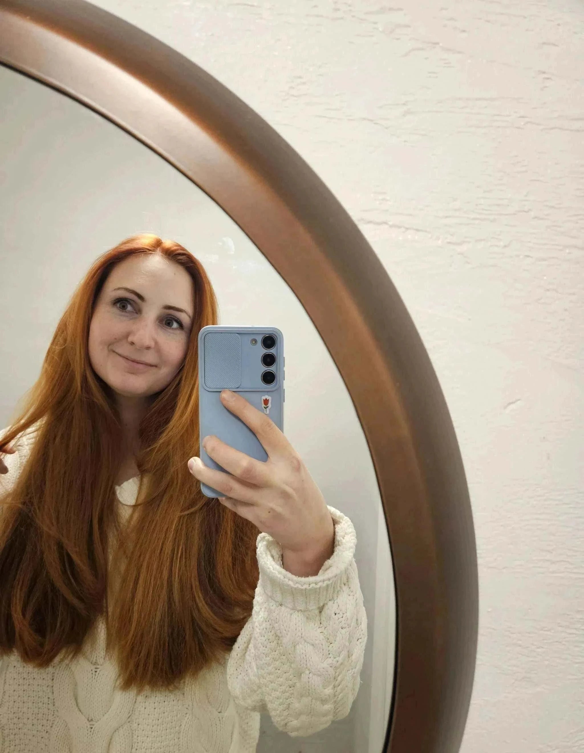 moteris ilgais raudonais plaukais daro nuotrauką veidrodžiuose su telefonu baltoje megztinėje
