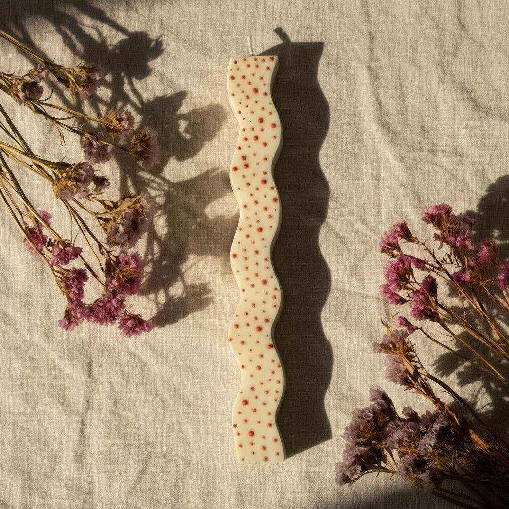 Dekoruota sojų vaško žvakė su raudonais tašeliais ir banguota forma ant audinio, šalia džiovintos gėlės