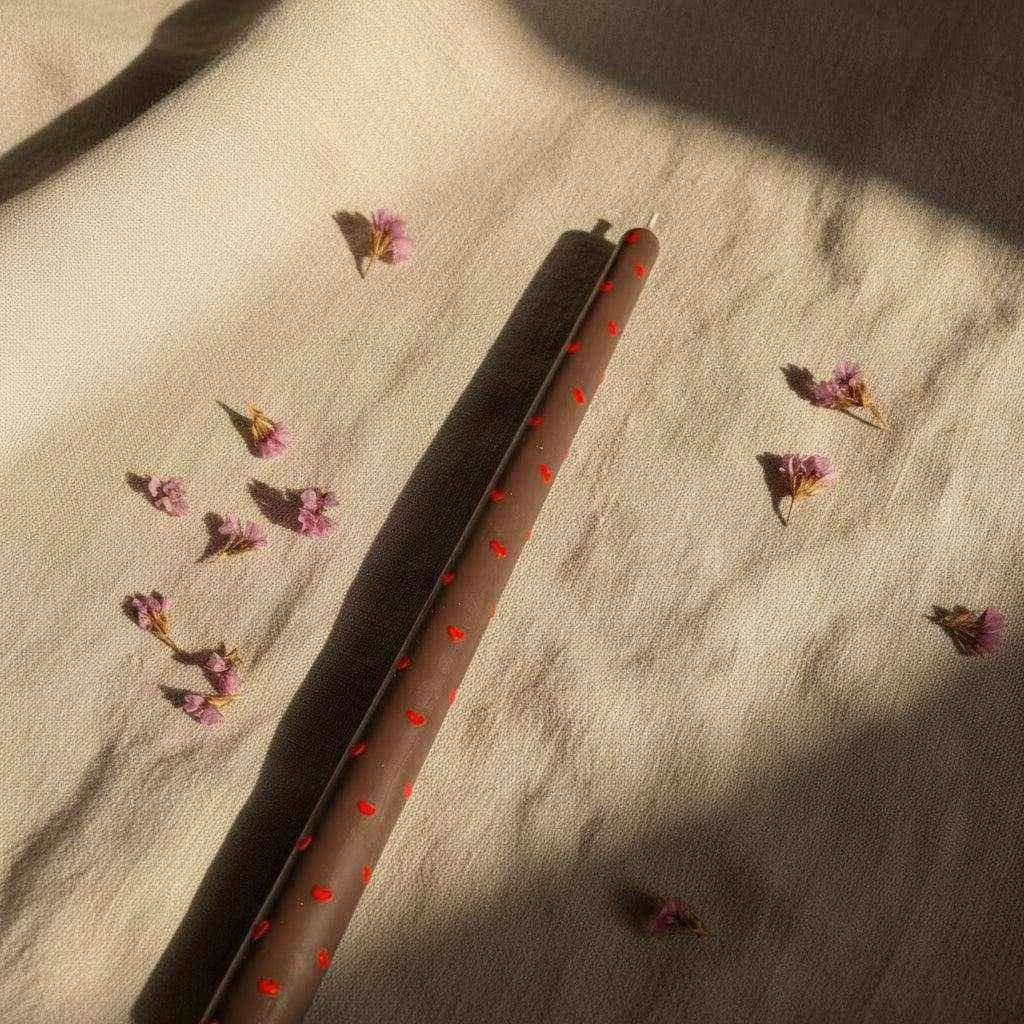 Dekoruota sojų vaško ilga plona žvakė su raudonais taškeliais ir rankų darbo elementais ant šviesaus audinio su mažais purpuriniais žiedais