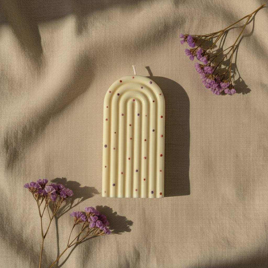 Dekoruota sojų vaško žvakė su smulkiais tašeliais ant natūralaus fono su violetinėmis gėlėmis
