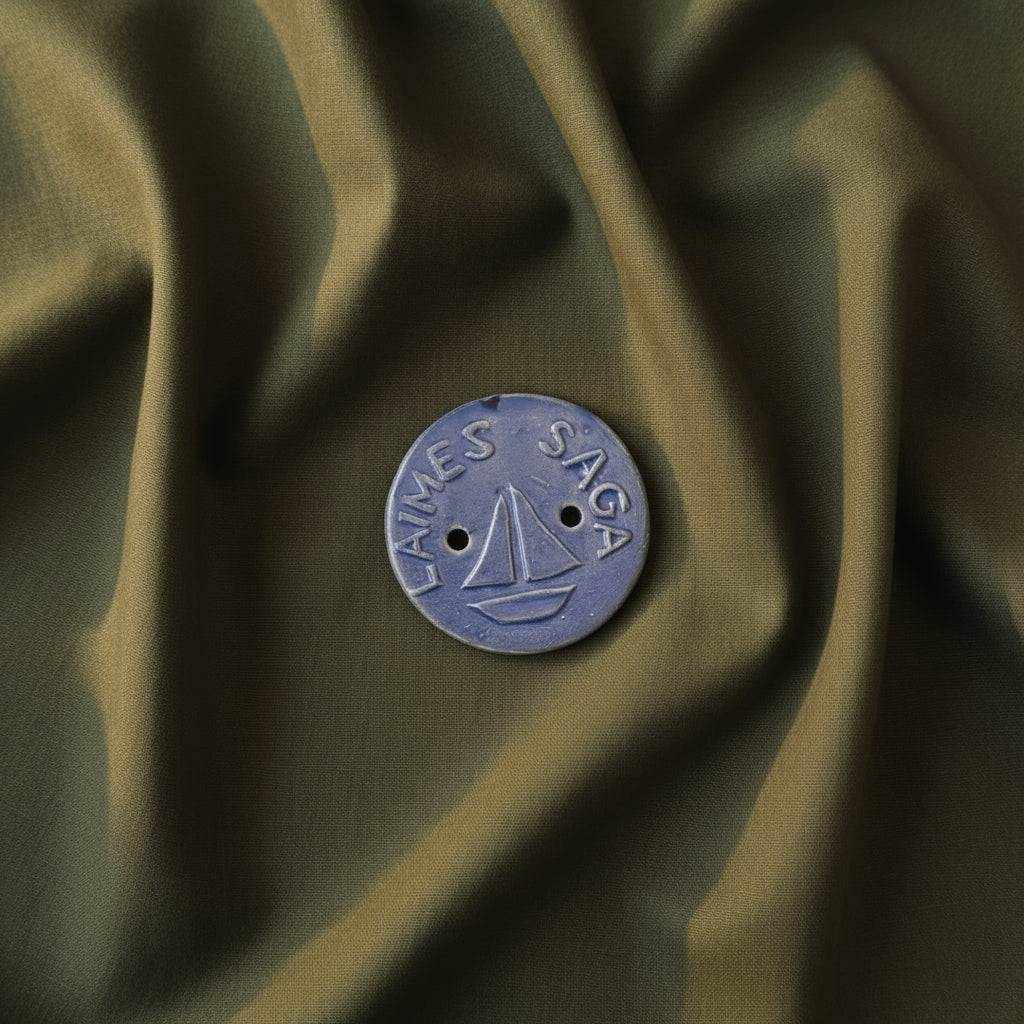 Rankų darbo keraminė Laimės saga su burlaivio simboliu ant tamsios medžiagos fono