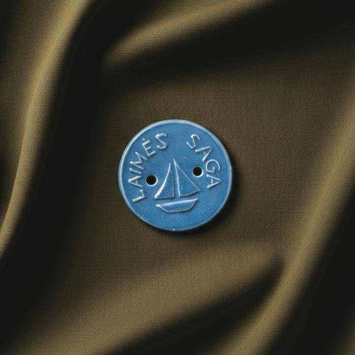 mėlyna keraminė Laimės saga su burlaivio simboliu ant rudos medžiagos fono